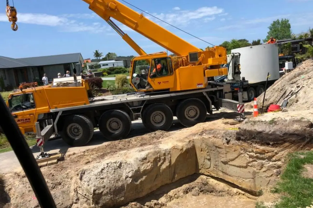 A large yellow mobile crane lifting heavy concrete tanks next to a deep site excavation managed by CB Earthmoving Northland.